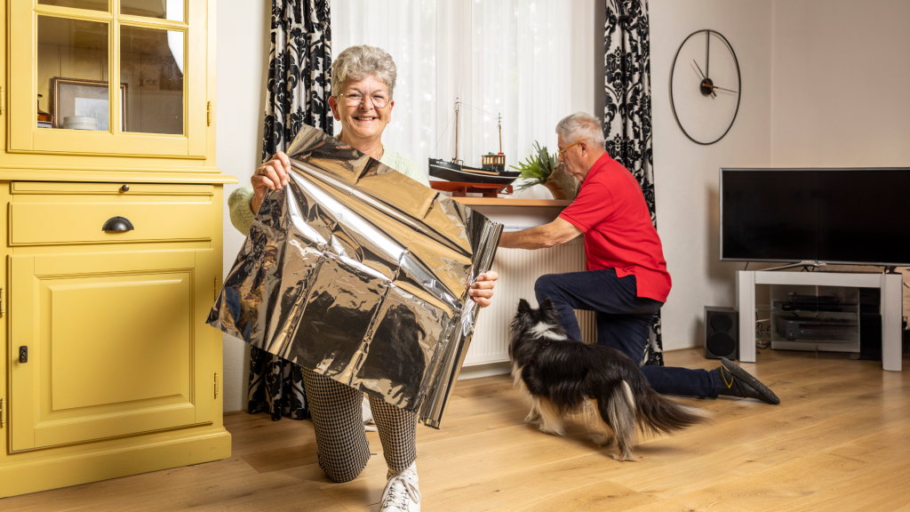 Een oudere vrouw toont isolatiefolie glimlachend aan de camera, terwijl een man op de achtergrond de folie aanbrengt achter een radiator.