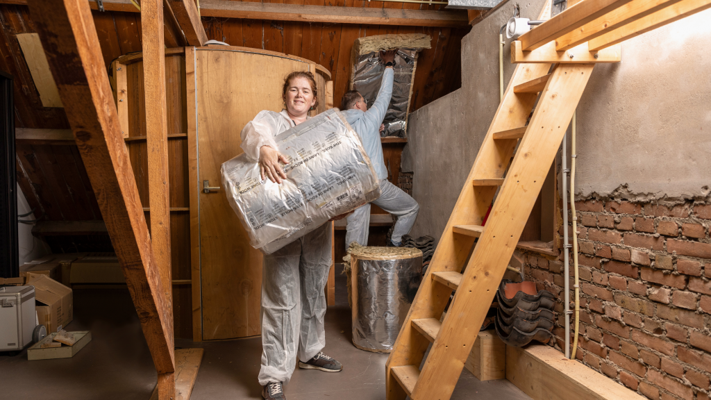 Twee mensen in witte overalls zijn bezig met het isoleren van een zolder. De persoon op de voorgrond houdt een grote rol isolatiemateriaal vast, terwijl de persoon op de achtergrond isolatiemateriaal aanbrengt aan een muur. De zolder heeft houten balken en een houten trap.