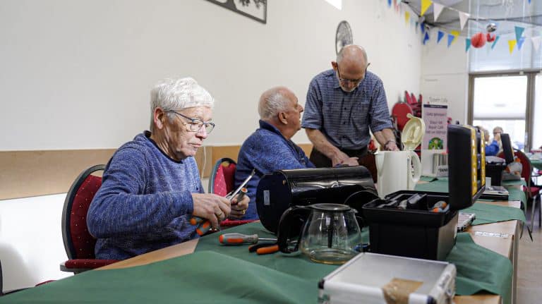 3 vrijwilligers van het repaircafé. Zij zitten aan een lange tafel. Daarop staat een gereedschapkist en verschillende apparaten. Waaronder een waterkoker. Twee vrijwilligers overleggen. Een vrijwilliger heeft schroevendraaiers in haar handen. (foto: Korver Fotografie)