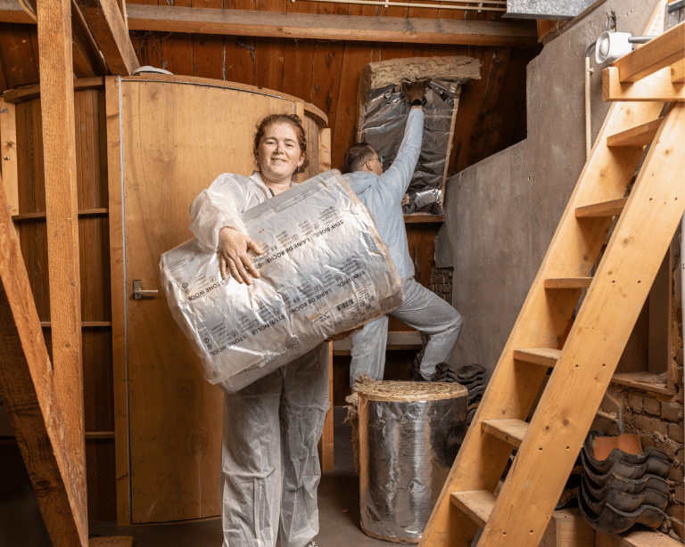 Twee personen werken op een zolder aan het aanbrengen van isolatie. Eén persoon houdt een grote rol isolatiemateriaal met reflecterende folie vast, terwijl de ander een stuk isolatie in het plafond plaatst. Beiden dragen beschermende overalls. De zolder heeft houten balken, een houten deur en een ladder die naar boven leidt.