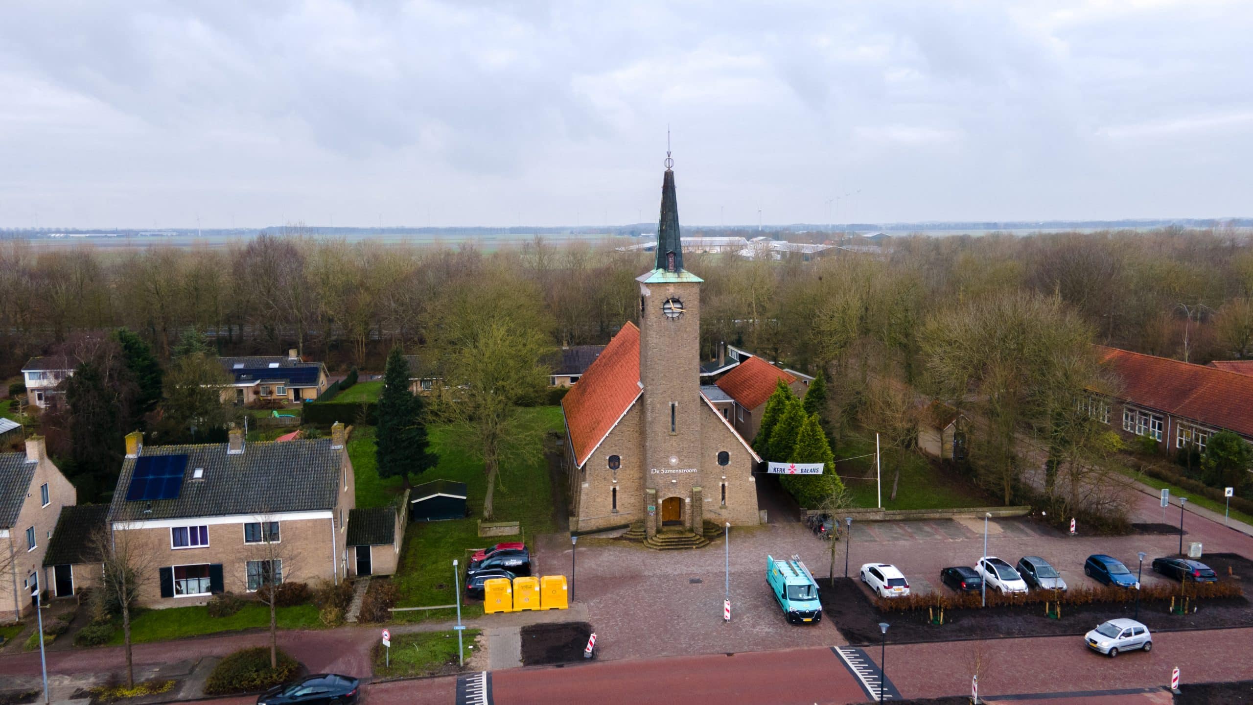 Luchtfoto van het gebouw van De Samenstroom: een kerkachtig pand met een hoge toren en klok, omringd door bomen. Er staan auto’s geparkeerd op het plein ervoor en er zijn woningen en groen in de omgeving te zien.