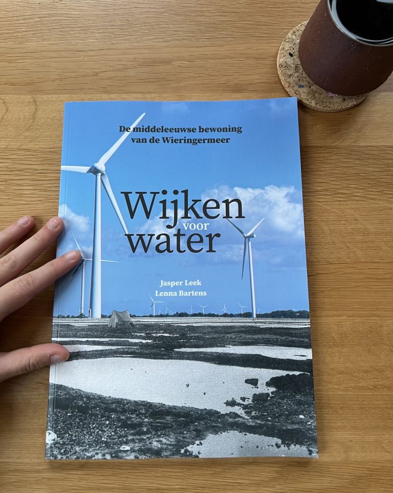 Boek met de titel ‘Wijken voor water – De middeleeuwse bewoning van de Wieringermeer’, met op de kaft een foto van windmolens in een open landschap. Een hand houdt het boek vast op een houten tafel.